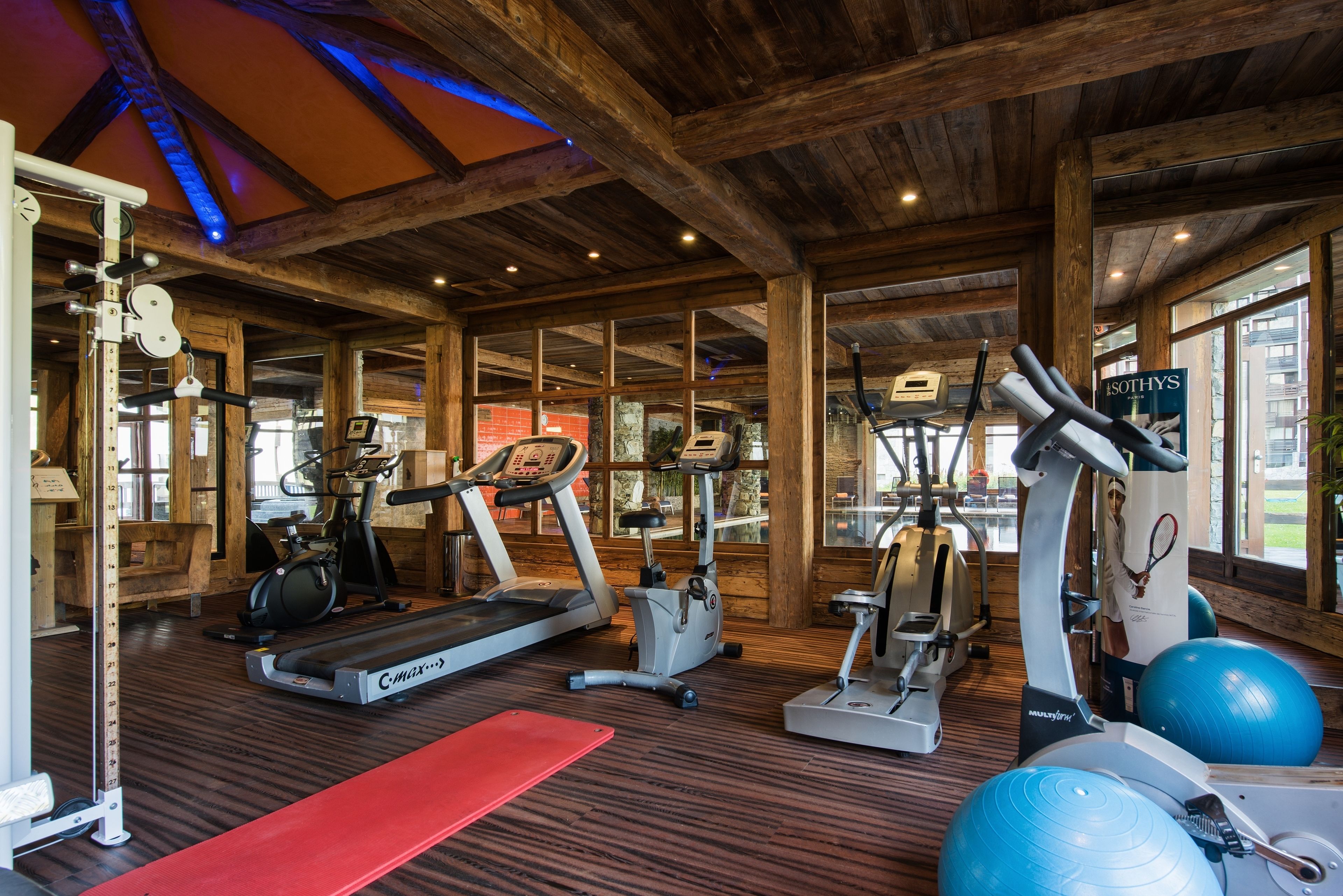 A gym with various exercise equipment, wooden beams on the ceiling, and large windows.