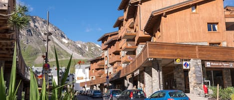 A wooden building with balconies, a sign for Maison de Tignes, and parked cars on a street with mountains in the background.