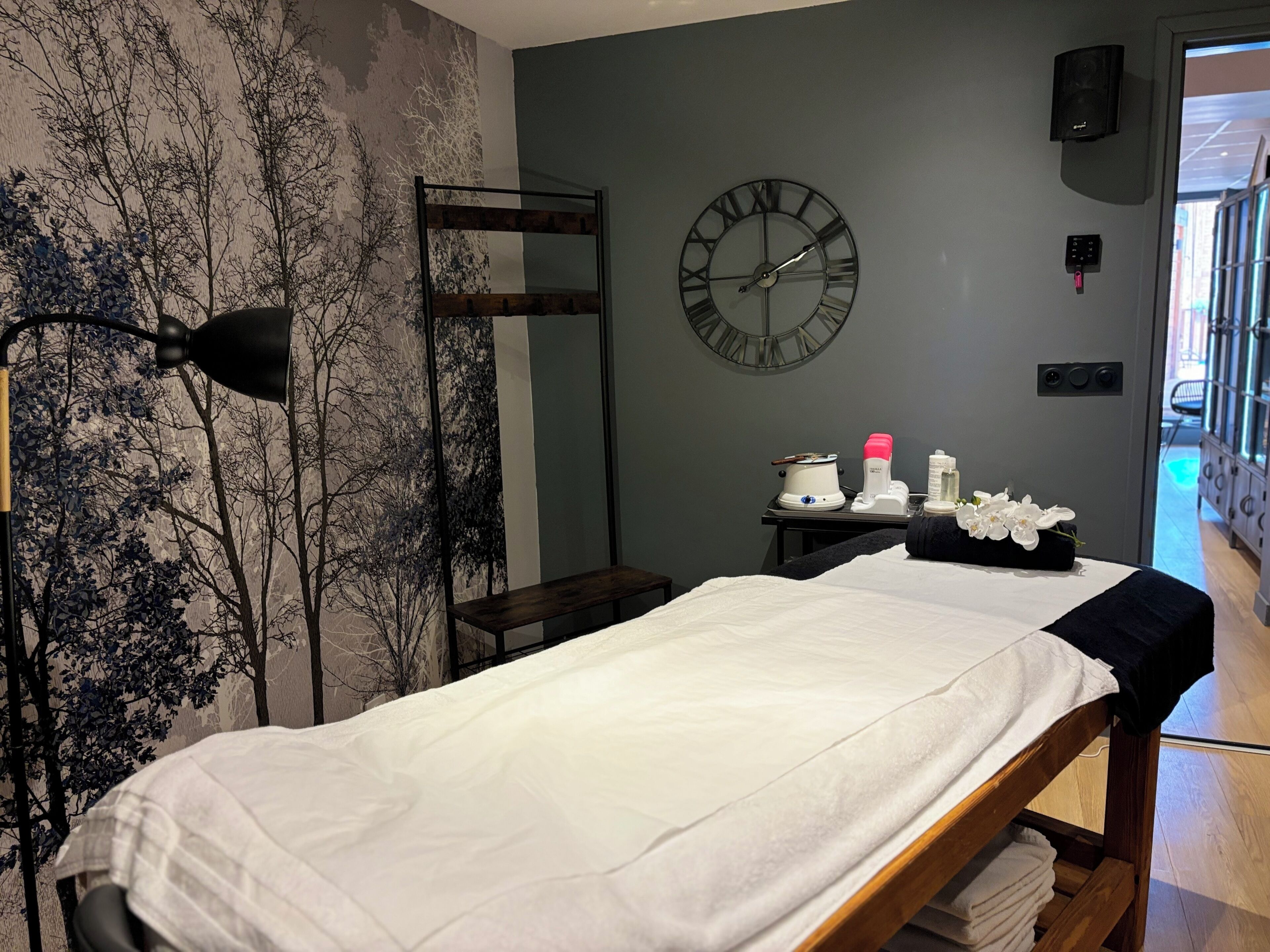 A room with a bed, a wall clock, a massage table, a massage chair, and a small table with bottles and a flower arrangement.
