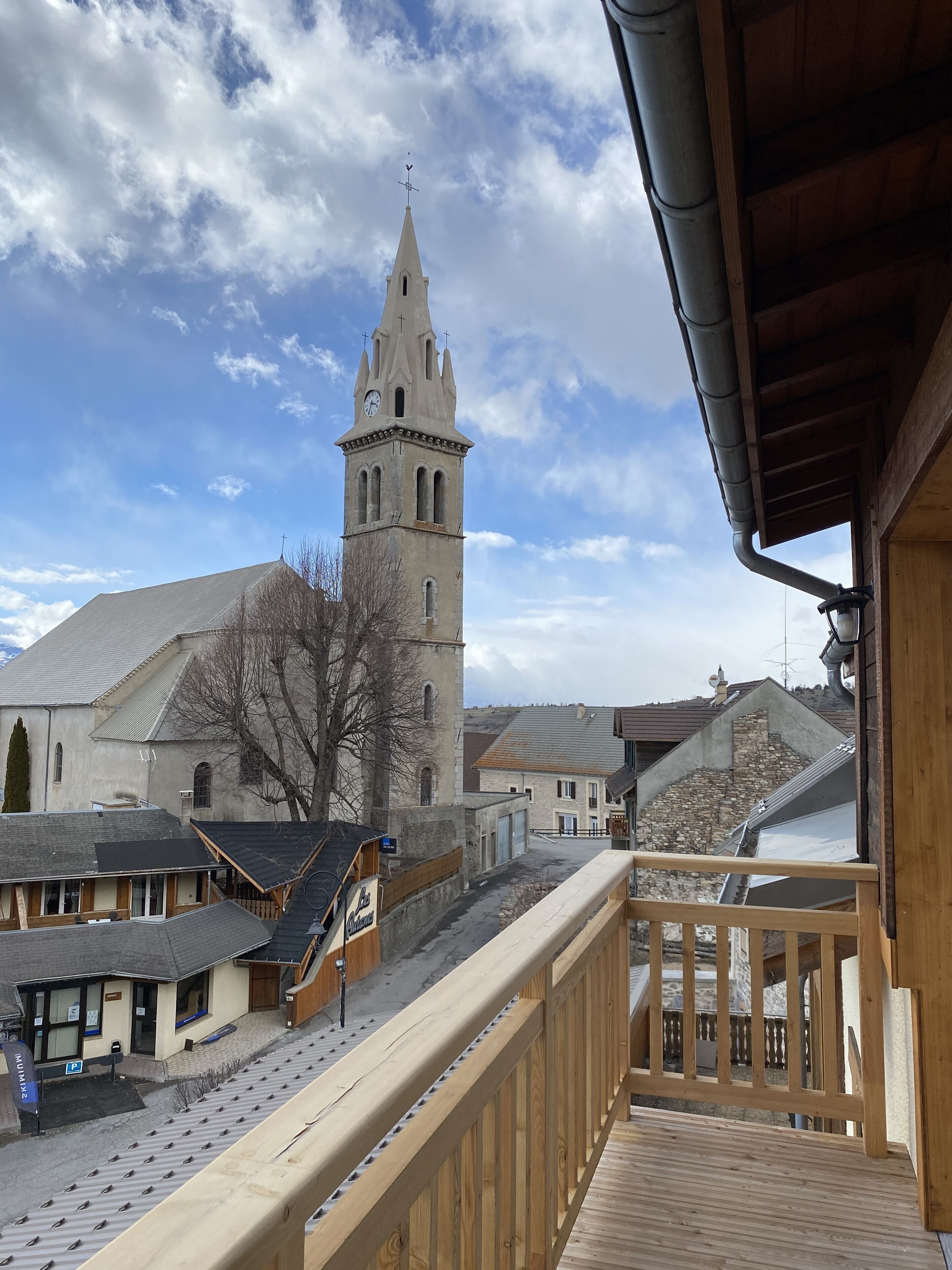 On voit une église dotée d’un haut clocher et d’un balcon en bois surplombant une rue bordée d’immeubles et d’une zone de stationnement.