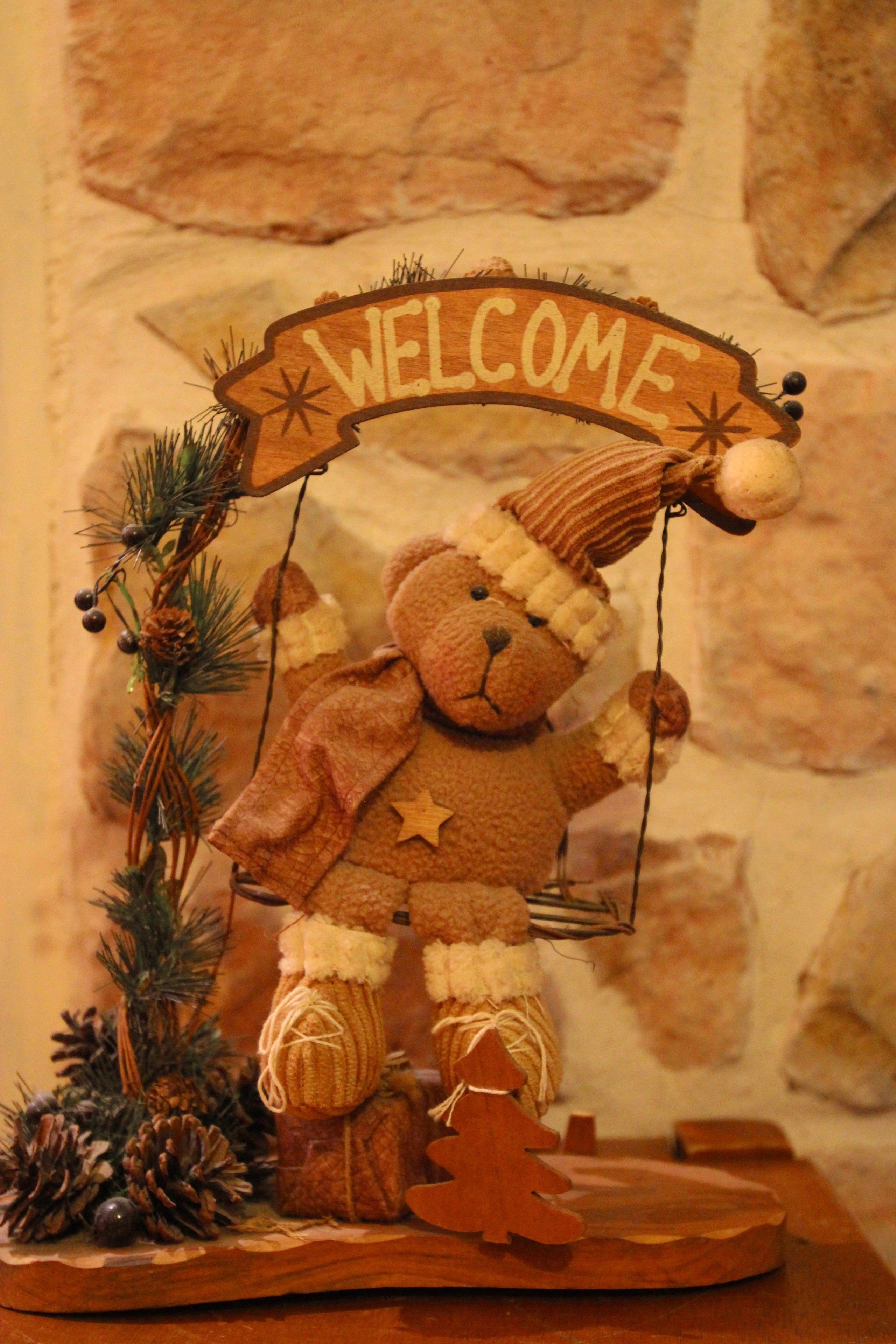 Un osito de peluche en un columpio con un letrero que dice 'WELCOME', rodeado de piñas y un pequeño árbol de Navidad.