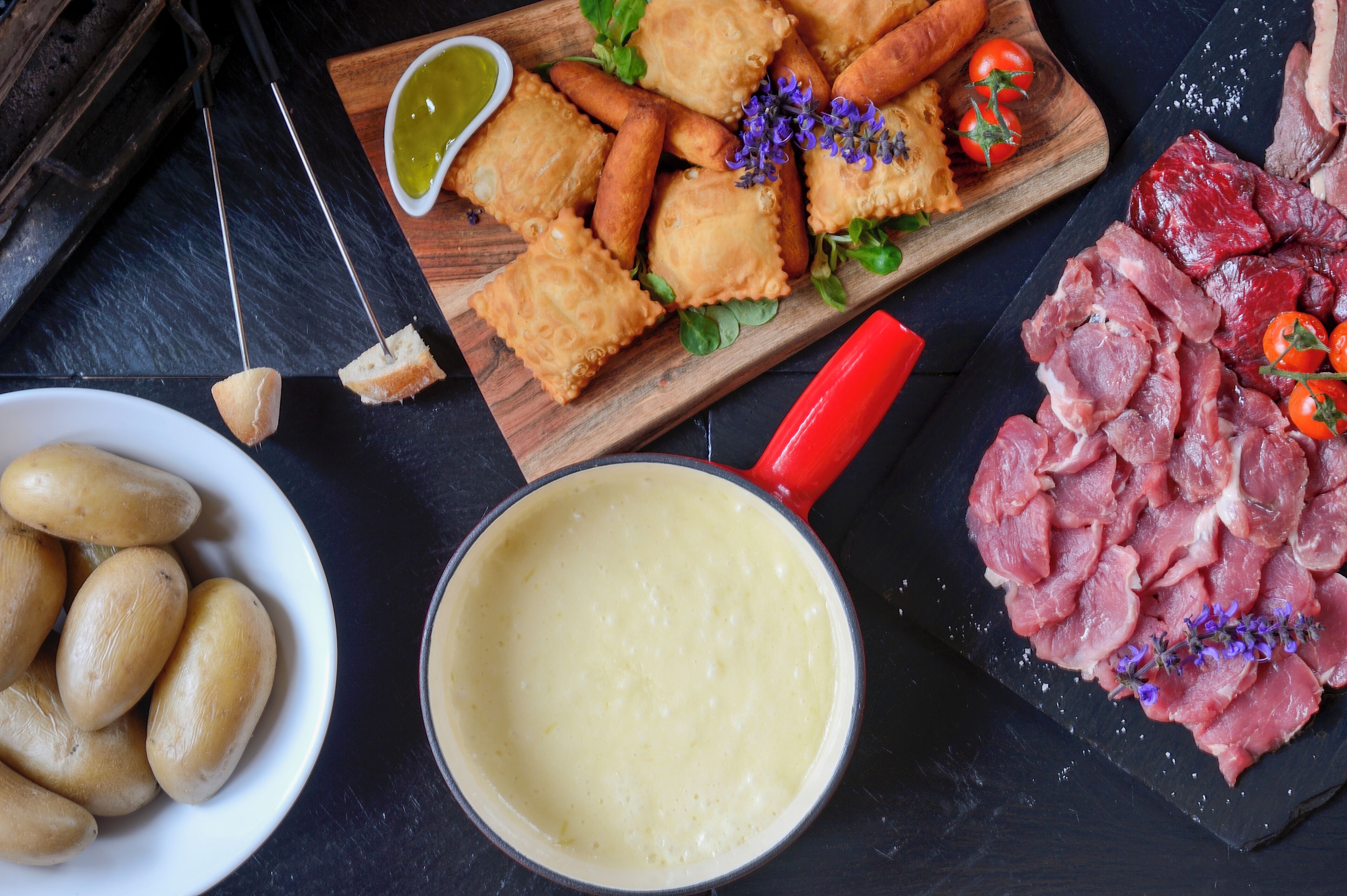 A variety of food items including fried pastries, a bowl of potatoes, a pot of sauce, and a platter of sliced meat, all arranged on a dark surface.