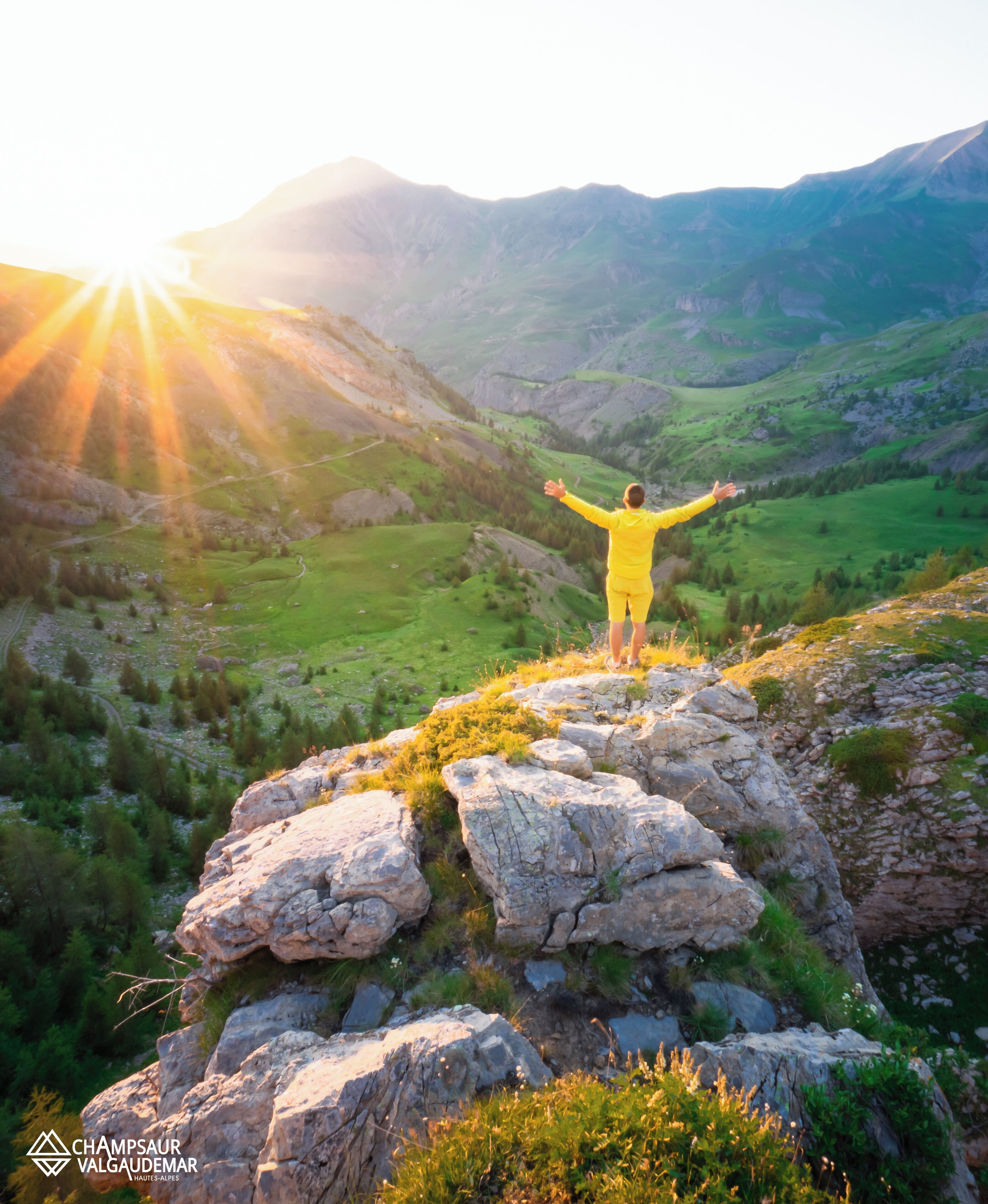 A person in a yellow outfit standing on a rocky outcrop with arms outstretched, overlooking a mountainous landscape with a bright sun in the background.