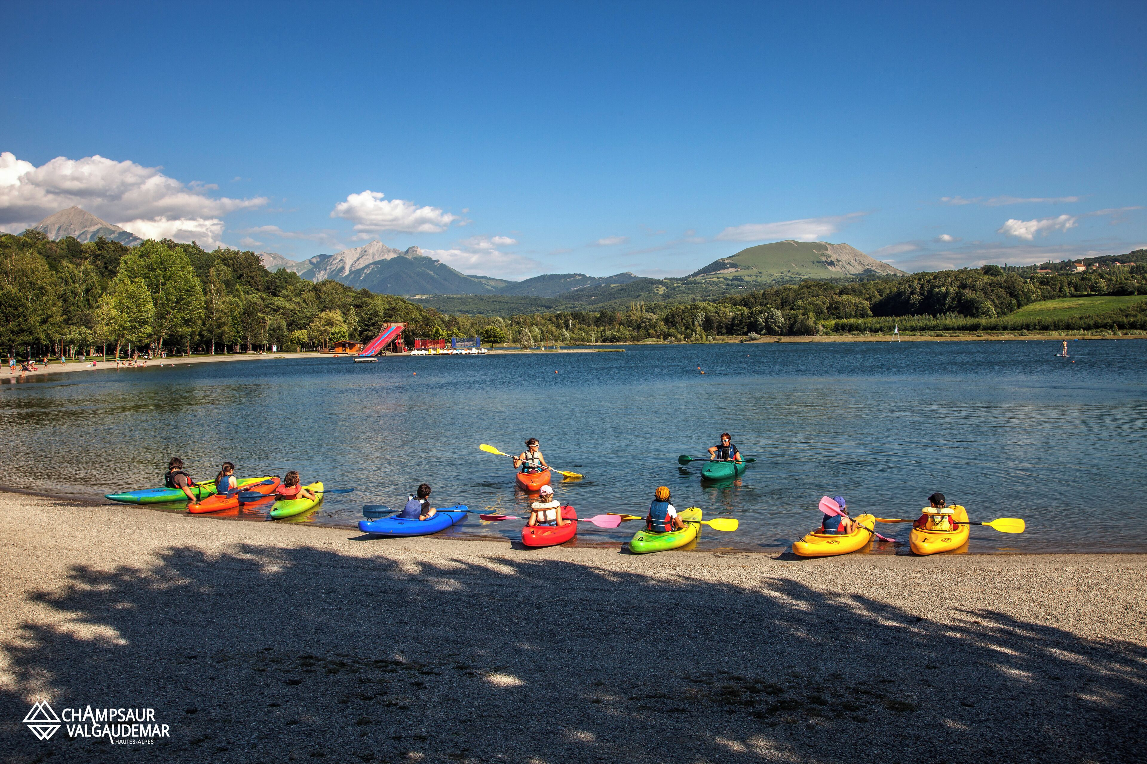 A group of kayaks lined up on a pebbly shore with a backdrop of mountains and a clear sky.