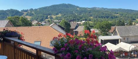 A balcony with a wooden railing overlooking a village with red-tiled roofs and a mountainous backdrop.