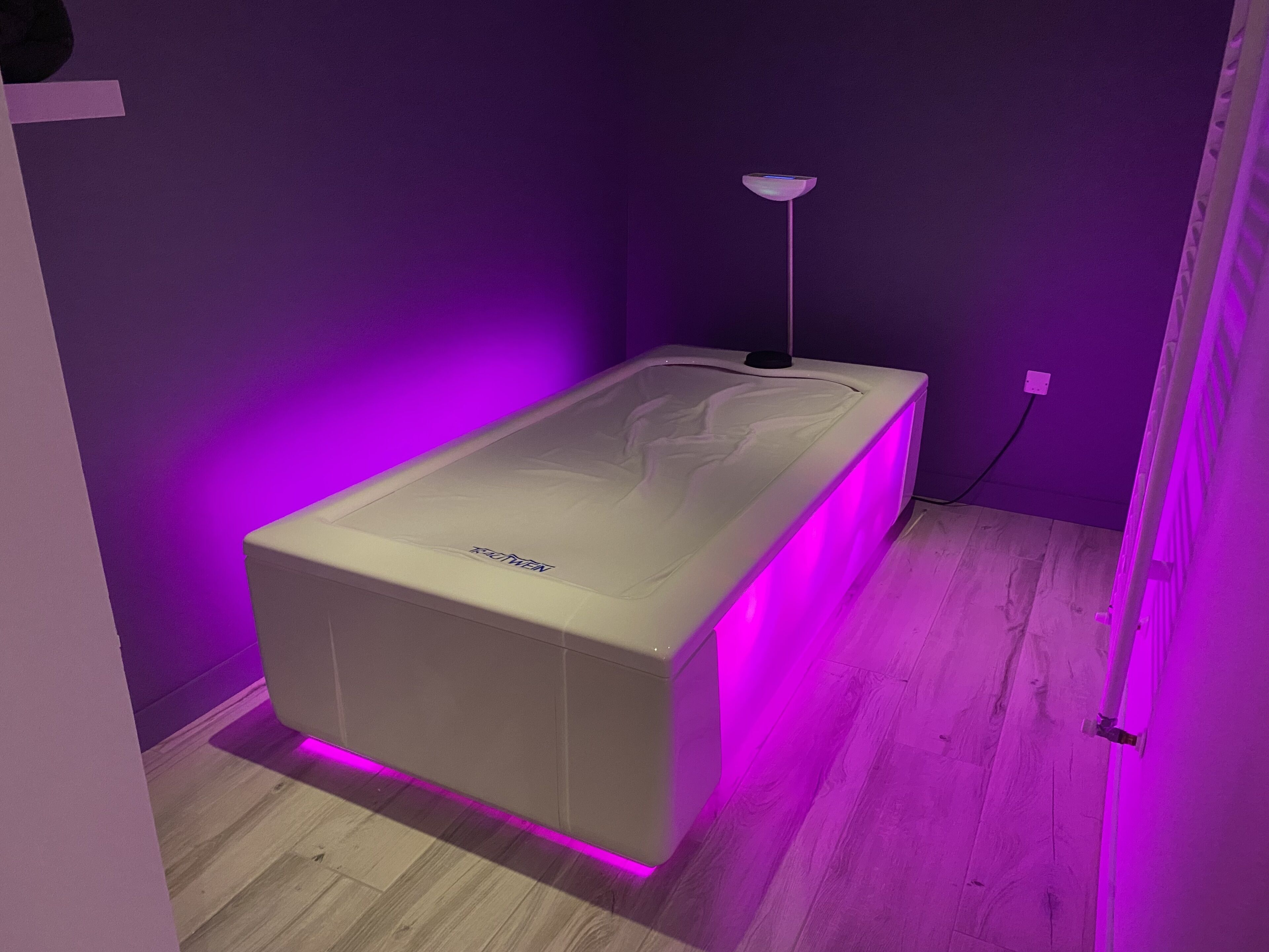 Ein Zimmer mit einem weißen Massagetisch, violetter Beleuchtung und Holzboden.