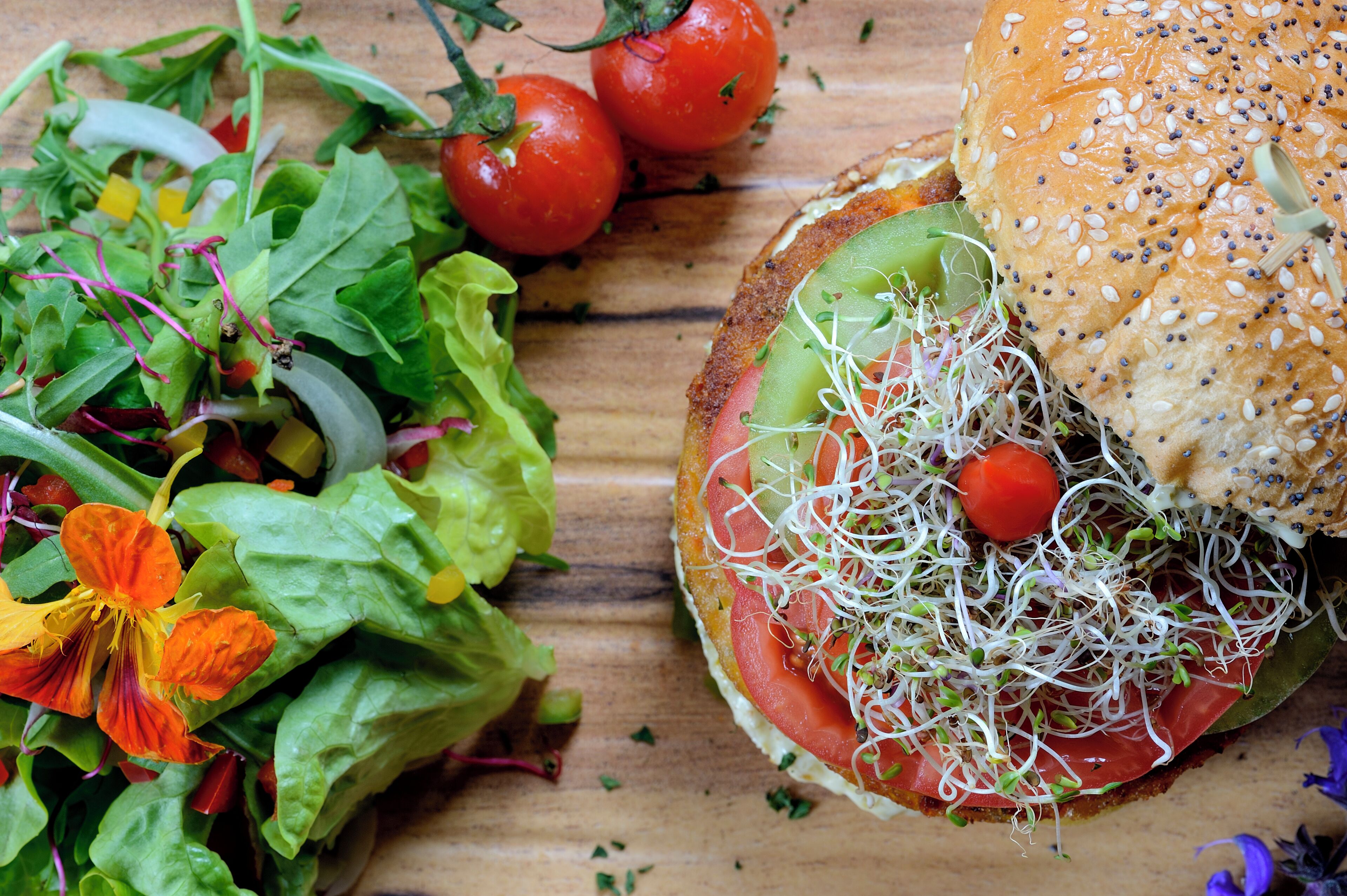 Un sándwich con lechuga, tomate y brotes sobre un pan, junto con una ensalada de diversas hojas verdes y una flor.