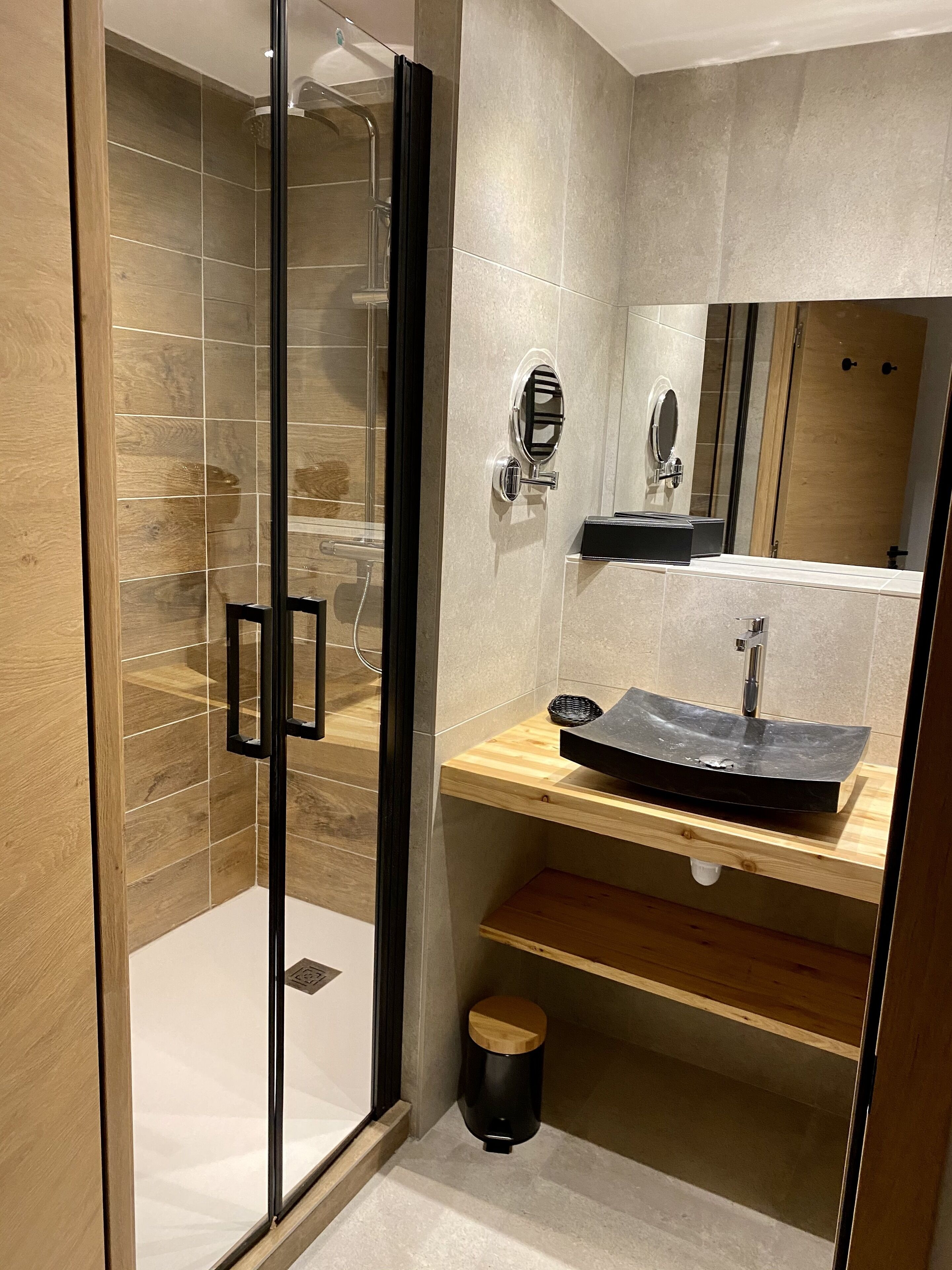 Un baño moderno con ducha envuelta en vidrio, tocador de madera con lavabo negro y espejo sobre el lavabo.