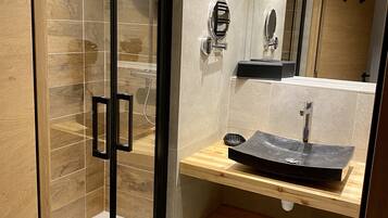 Un baño moderno con ducha envuelta en vidrio, tocador de madera con lavabo negro y espejo sobre el lavabo.