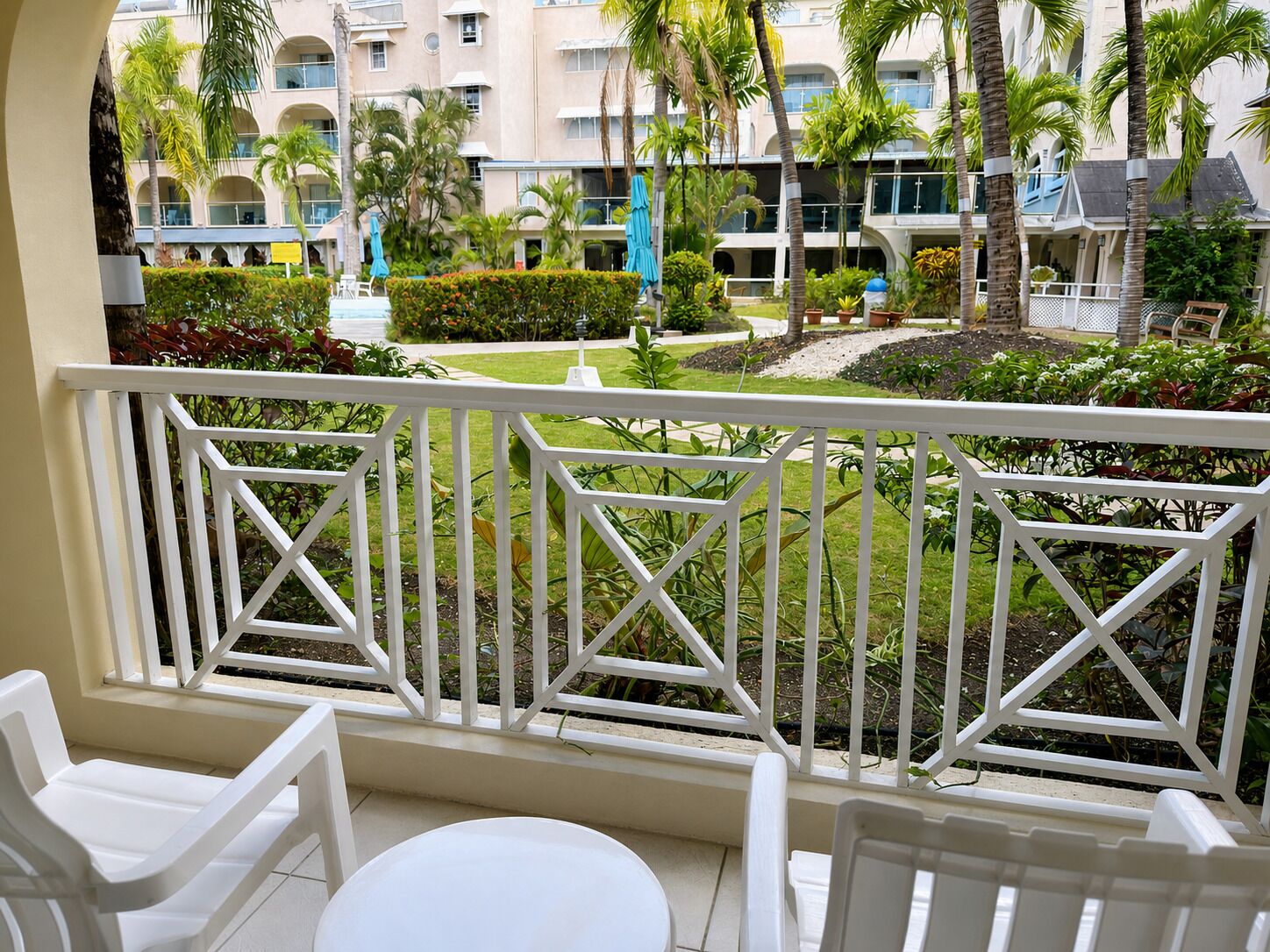A balcony with white railings, a white table, and white chairs. In the background, there is a building with multiple balconies and palm trees.