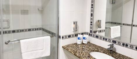Un baño con cabina de ducha de vidrio, un lavabo blanco con encimera de granito y un espejo grande con un marco decorativo.