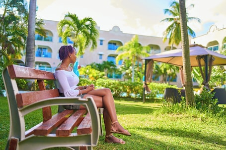 A person sitting on a wooden bench in a lush, green outdoor area with palm trees and a building in the background.
