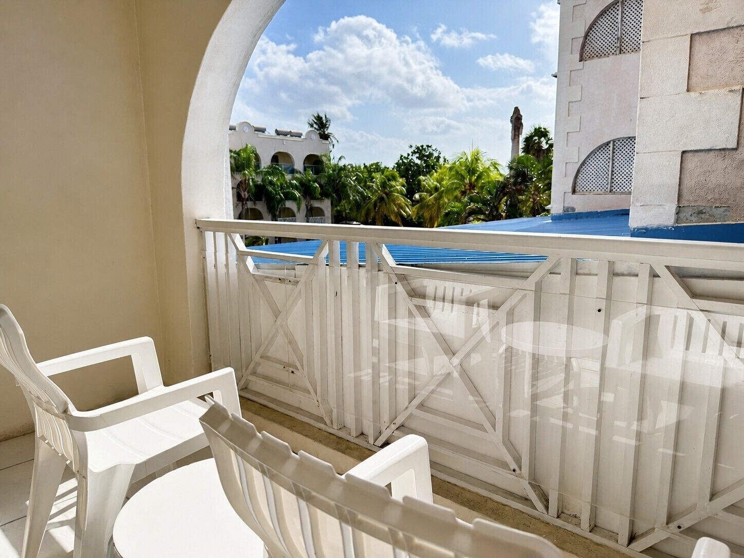 Un balcon avec des chaises en plastique blanches et une balustrade blanche, donnant sur une piscine et un bâtiment orné d’arches.