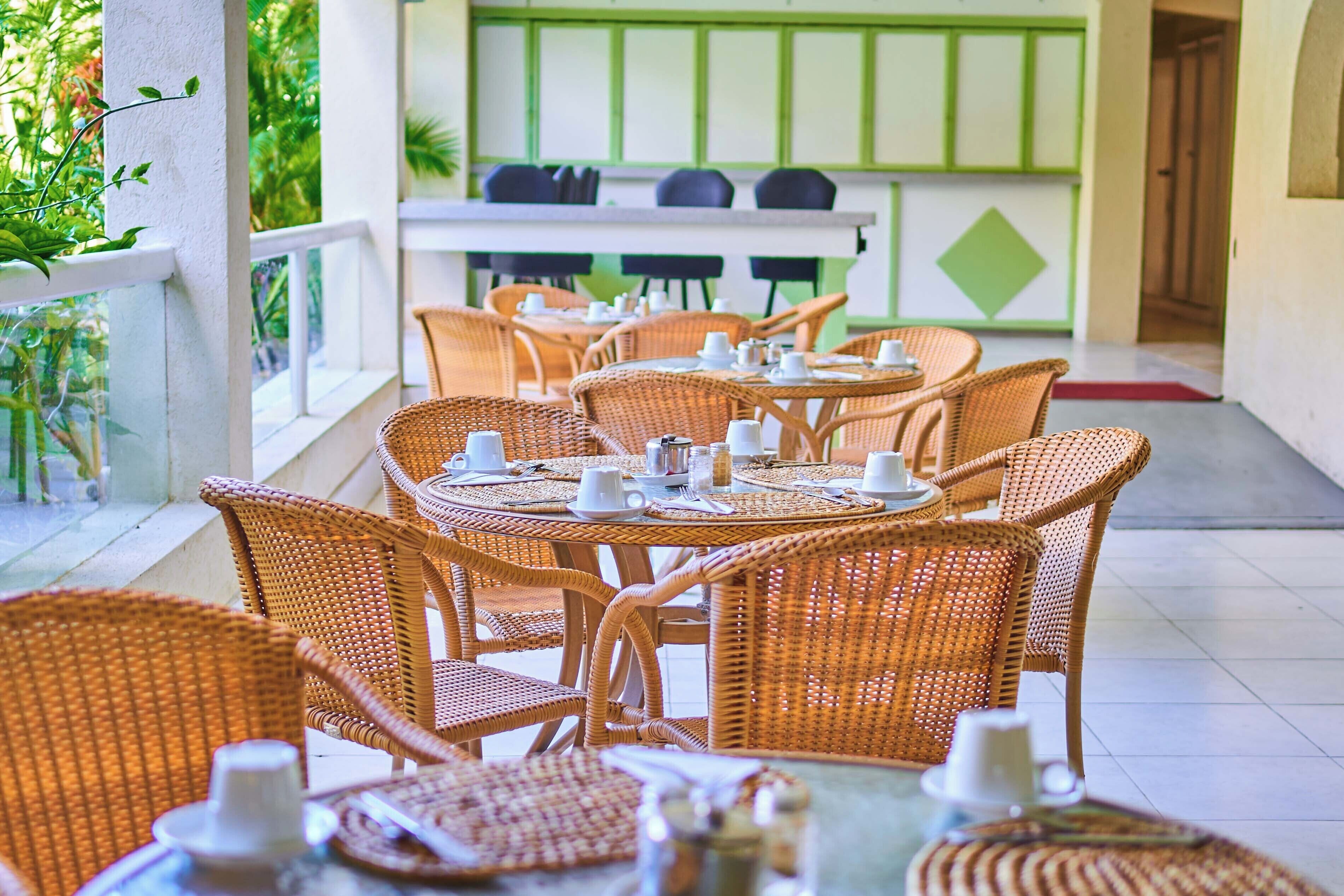 A restaurant with wicker tables and chairs, a counter with kitchen appliances, and a view of greenery outside.