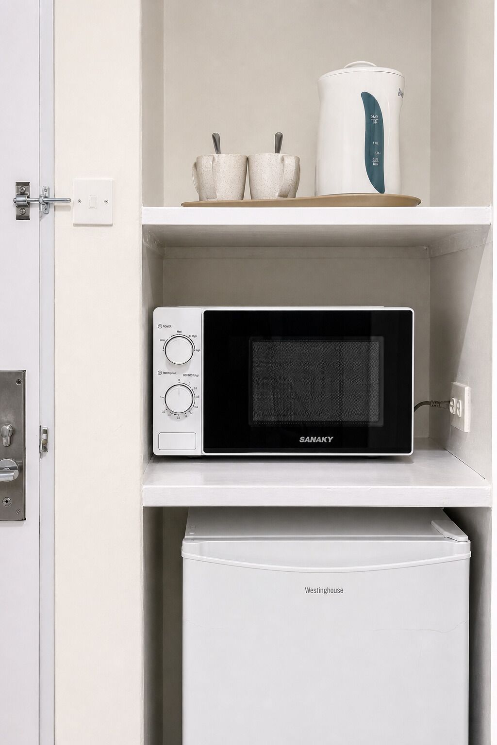 A microwave oven with a digital display and control knobs, a white kettle, and two ceramic cups on a white shelf.