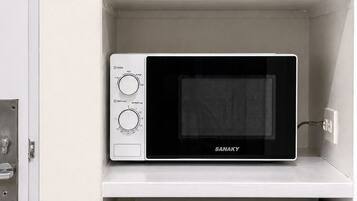 A microwave oven with a digital display and control knobs, a white kettle, and two ceramic cups on a white shelf.