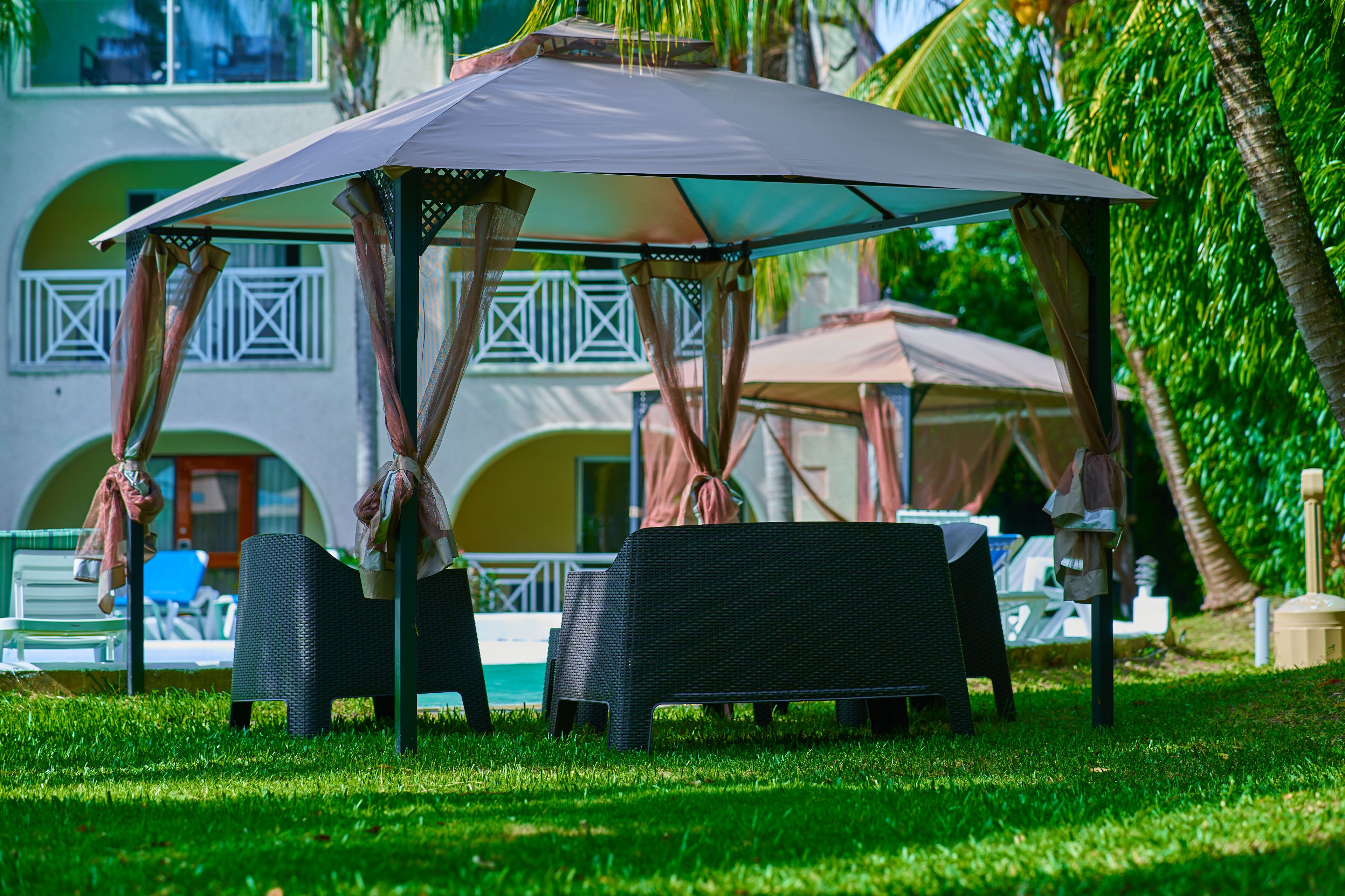 A gazebo with a table and chairs, surrounded by a pool and palm trees.