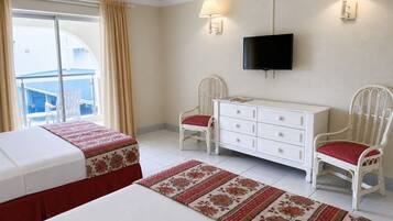 A hotel room with a bed, a dresser, two chairs, a television, and a view of the sea through a balcony.