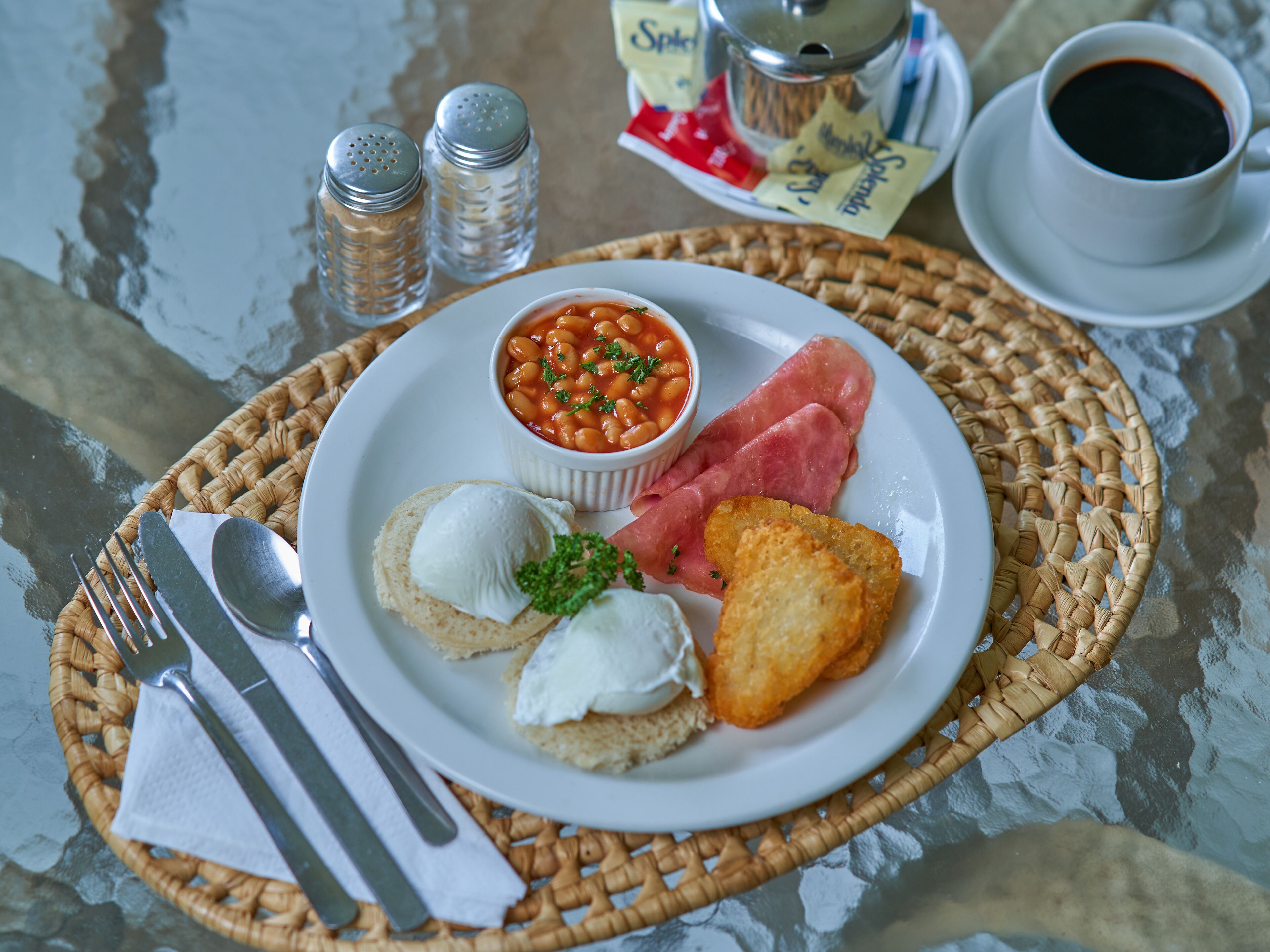 Un piatto con fagioli, prosciutto, pane tostato e una tazza di caffè su un tavolo con saliera e pepiera.