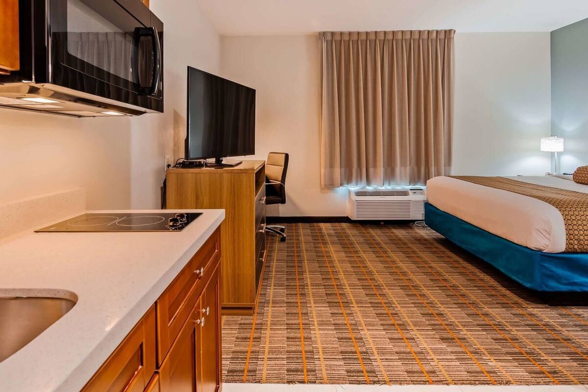 A hotel room with a kitchenette, a bed, a TV, and an air conditioning unit.