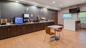 Zona cuina moderna amb cafetera, refrigerador i un dispensador de begudes. Hi ha una taula blanca amb dues cadires i una petita zona de bar amb tamborets.