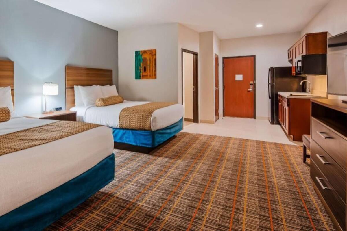 A hotel room with two beds, a kitchenette, a television, and a door leading to another room.