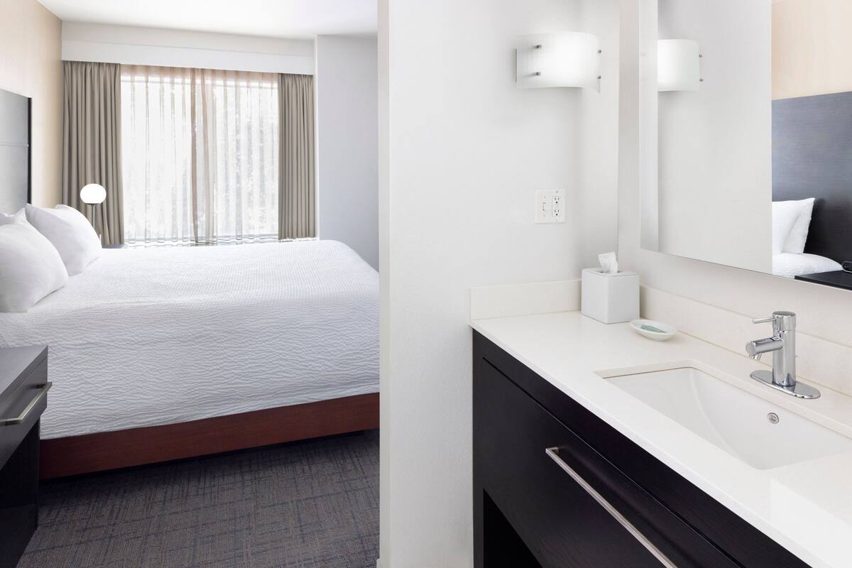 Une chambre d’hôtel comprenant un lit, une table de chevet, une commode, une grande fenêtre avec des rideaux, une salle de bain avec un lavabo, un miroir et un porte-serviettes.