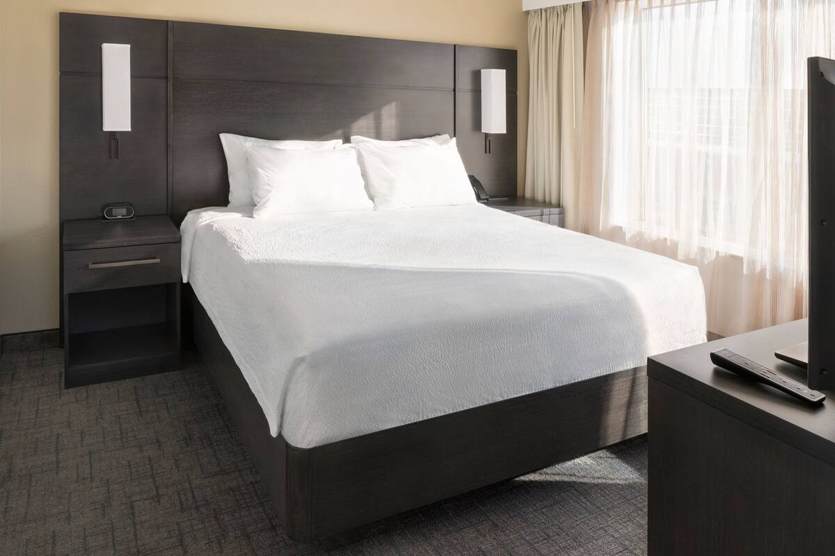 Une chambre d’hôtel moderne avec un grand lit, une table de chevet, une commode, une télévision et une fenêtre avec des rideaux.
