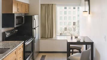 A compact kitchen with a microwave, oven, and refrigerator. A window with curtains provides natural light, and a table with chairs is positioned near the window.