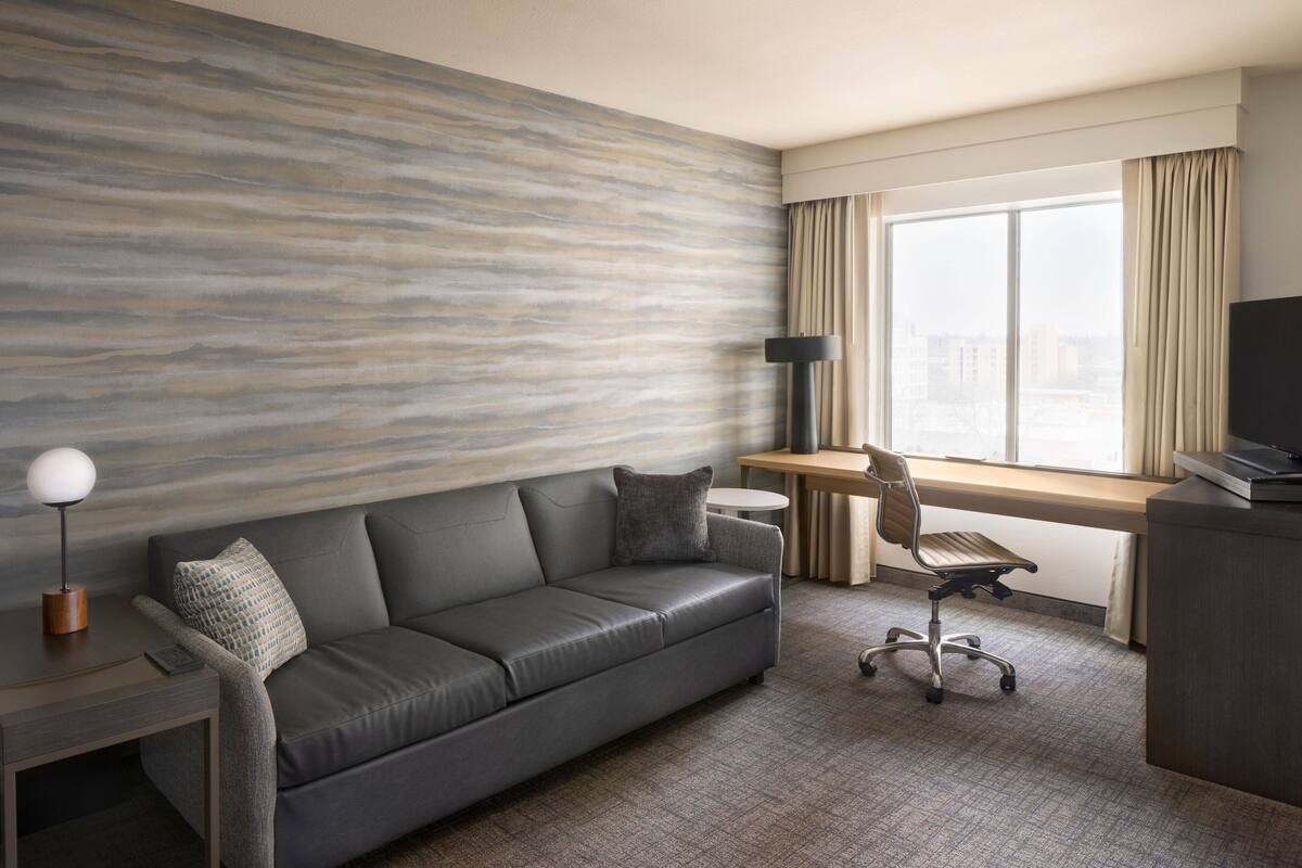 Une chambre d’hôtel moderne avec un canapé gris, un bureau avec une chaise, un téléviseur à écran plat et une grande fenêtre avec des rideaux.