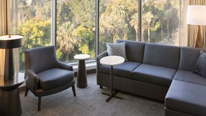 Un salon moderne comprenant un canapé, un fauteuil et des tables rondes, avec une grande fenêtre offrant une vue sur la ville et un parc.