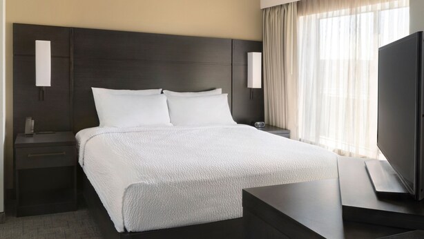 Ein modernes Hotelzimmer mit einem großen Bett, einem dunklen Kopfteil, Nachttischen, einem Flachbildfernseher und einem Fenster mit durchsichtigen Vorhängen.