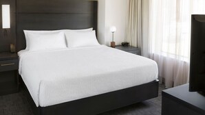 Un lit bien fait, avec du linge de lit blanc, une tête de lit sombre, une table de chevet avec une lampe et un téléviseur posé sur un meuble.