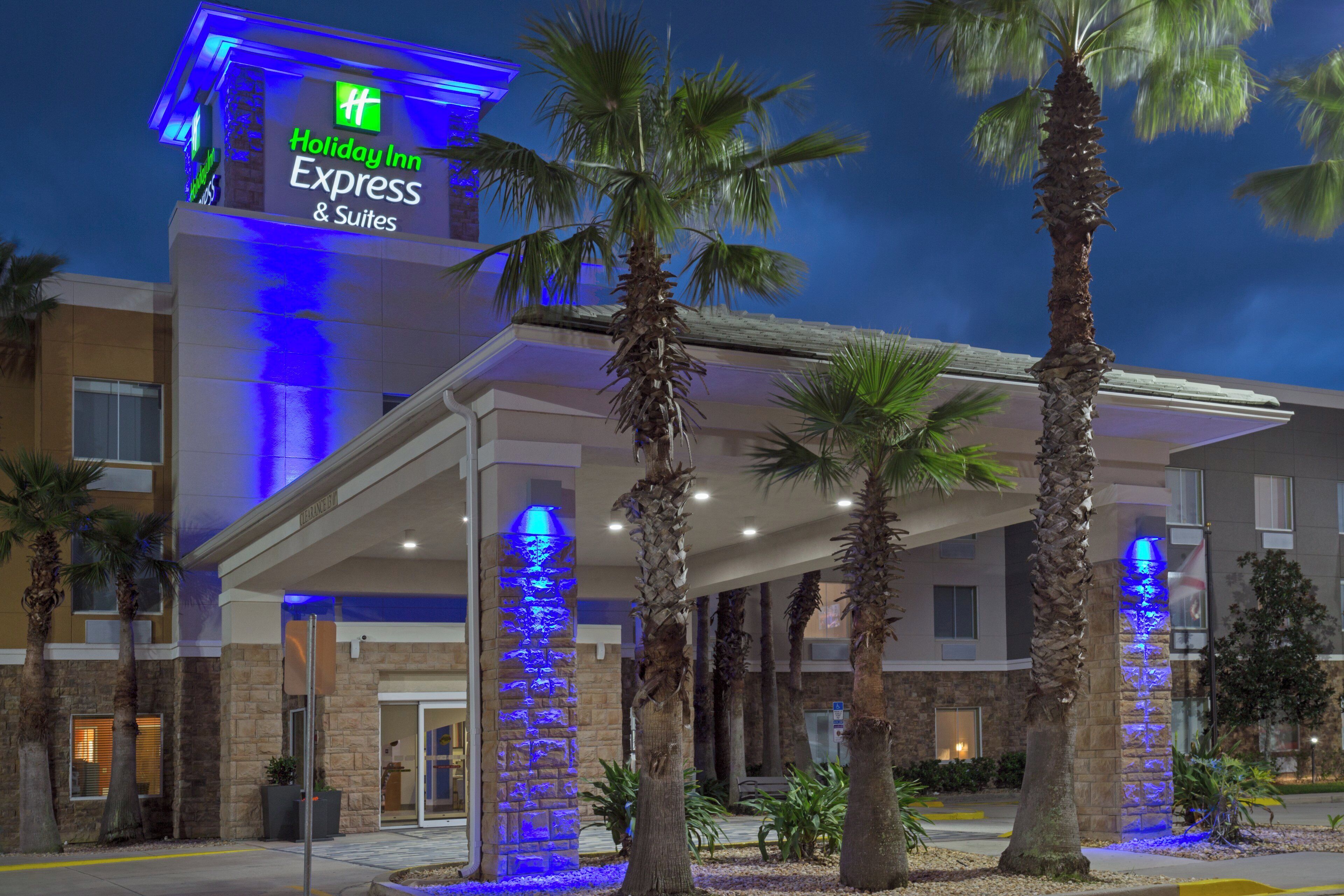 A Holiday Inn Express & Suites building with a covered entrance, palm trees, and a blue-lit sign.