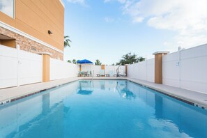 Une piscine à l’eau d’un bleu cristallin, entourée de murs blancs et d’un bâtiment en briques. On aperçoit des chaises longues et un parasol bleu à l’arrière-plan.
