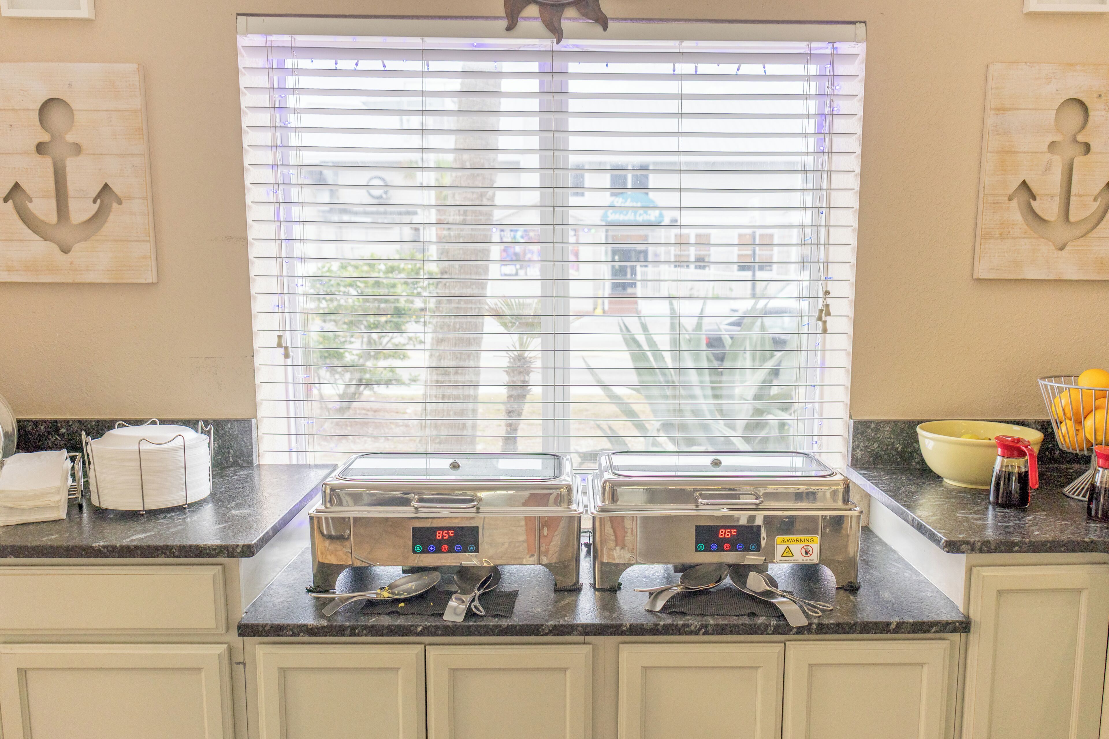 A kitchen with two stainless steel appliances, a window with blinds, and a bowl of fruit on the counter.
