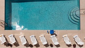Vista aérea de una piscina rectangular con agua azul cristalina, rodeada de sillas de descanso y una pequeña construcción azul.