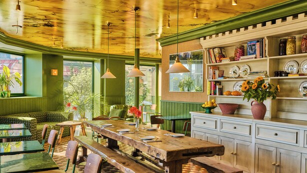 Un restaurante con una larga mesa de madera, bancos y una zona de cocina con estantes y frascos.