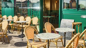 Un café al aire libre con sillas y mesas de mimbre, un toldo verde y un cartel de menú visible a través del ventanal.