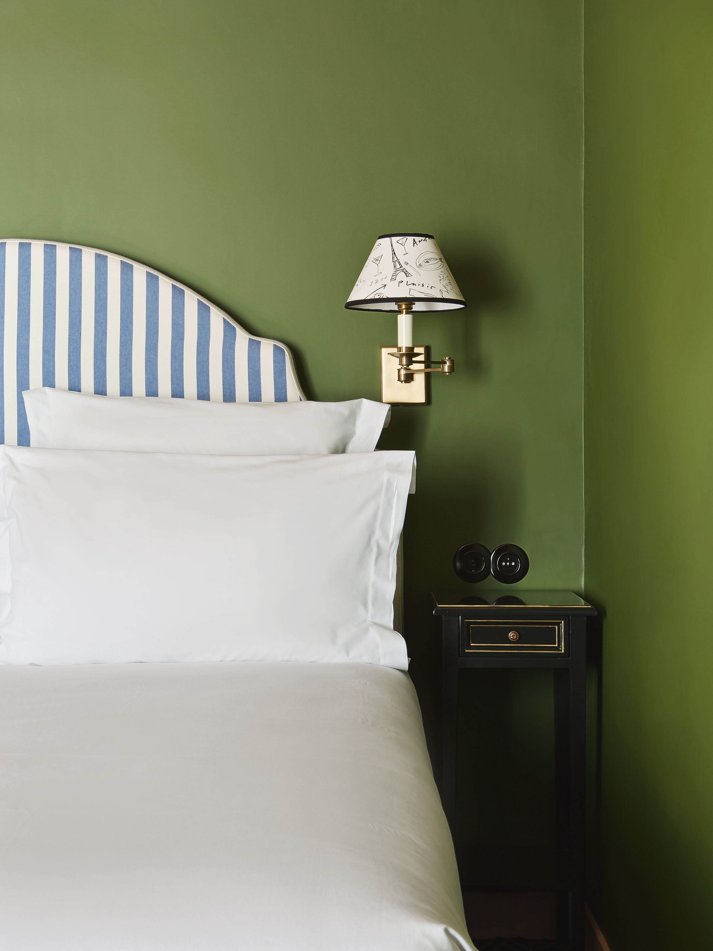 A bed with white linens and a striped headboard, a wall-mounted lamp with a patterned shade, and a small bedside table with a drawer.