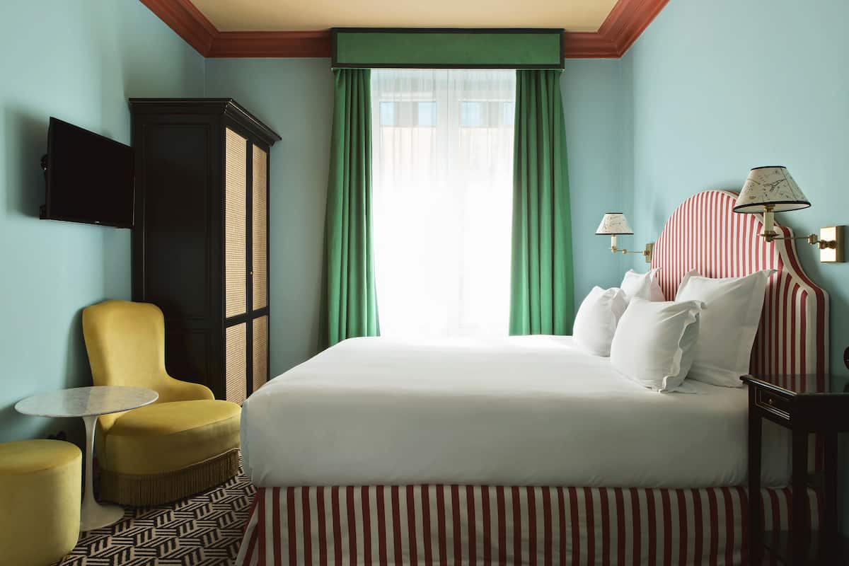 Une chambre d’hôtel avec un grand lit, un fauteuil jaune, une petite table ronde et un téléviseur fixé au mur.