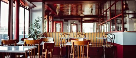 A restaurant interior with wooden chairs and tables, a patterned floor, and large windows allowing natural light.
