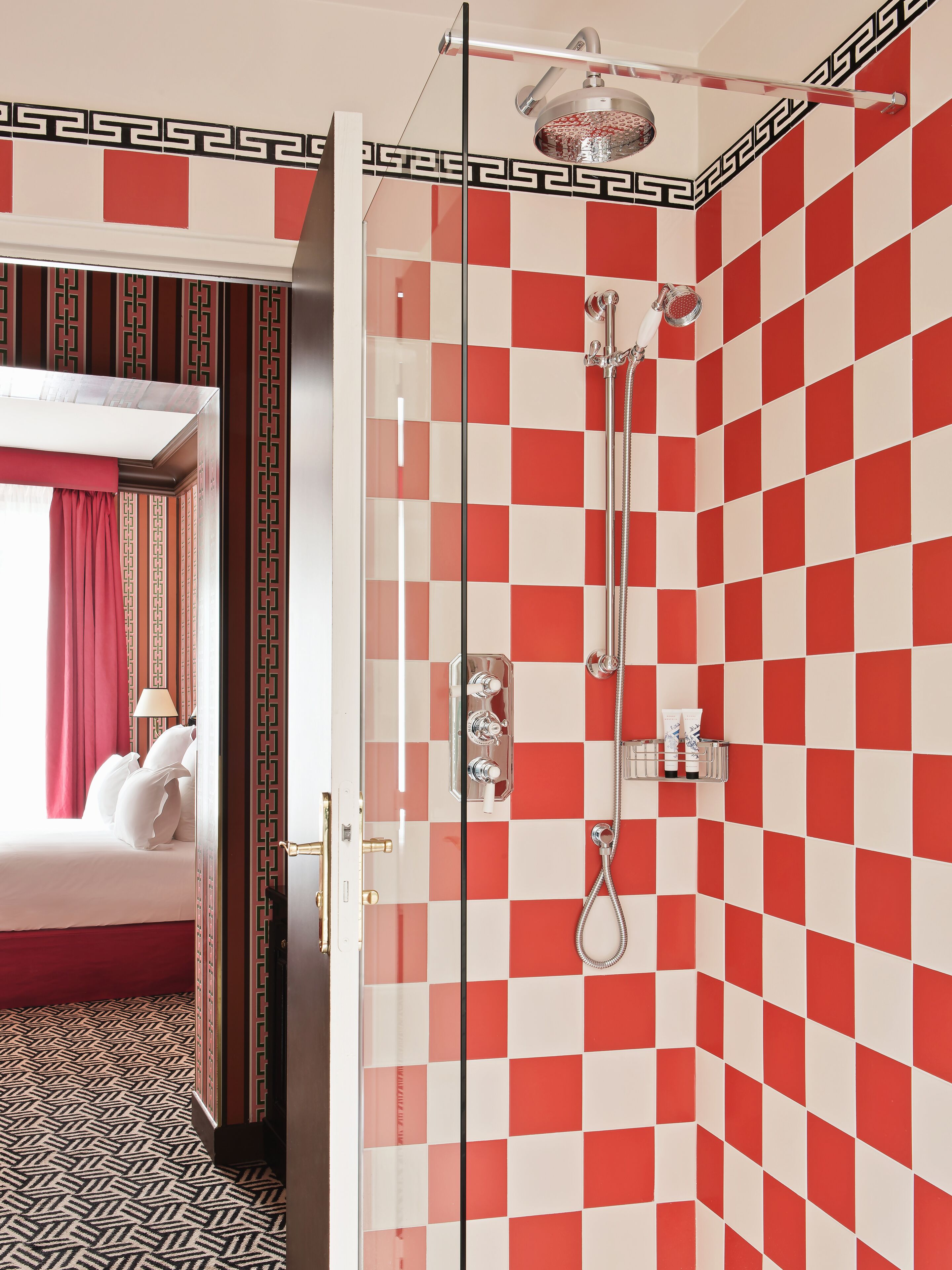 A hotel room with a checkered red and white wall, a shower with a glass door, and a bed with white linens.
