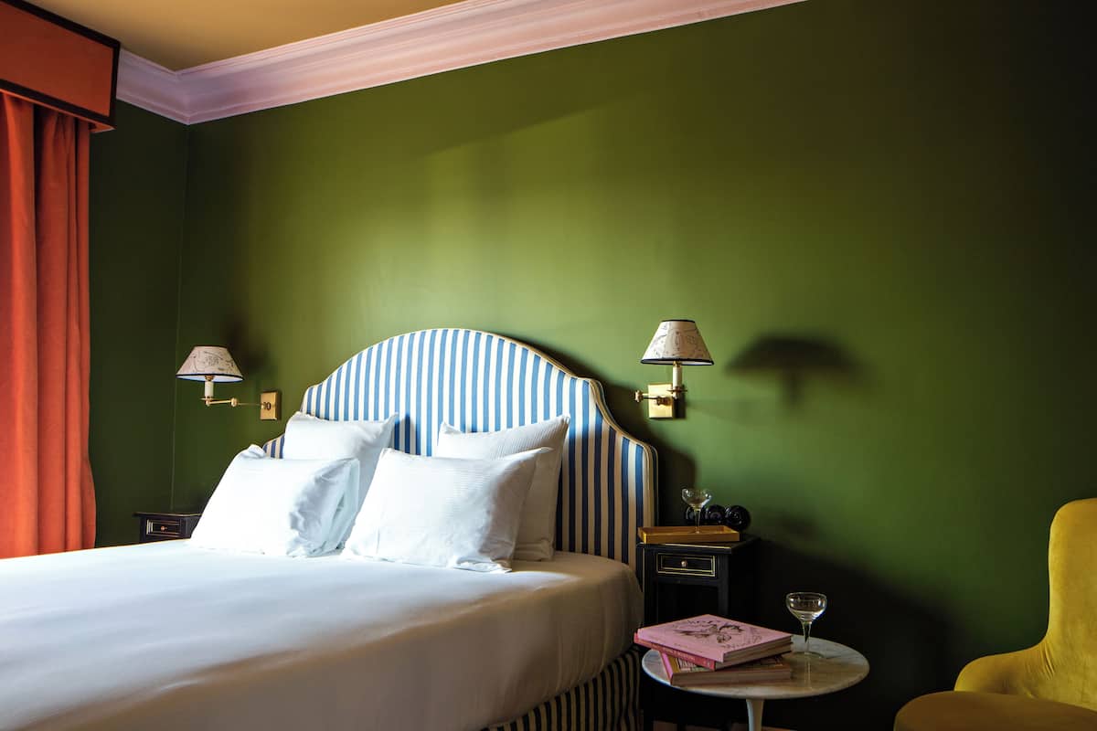 Une chambre d’hôtel avec un lit, une table de chevet avec une lampe, une chaise et une petite table sur laquelle se trouvent un livre et un verre.