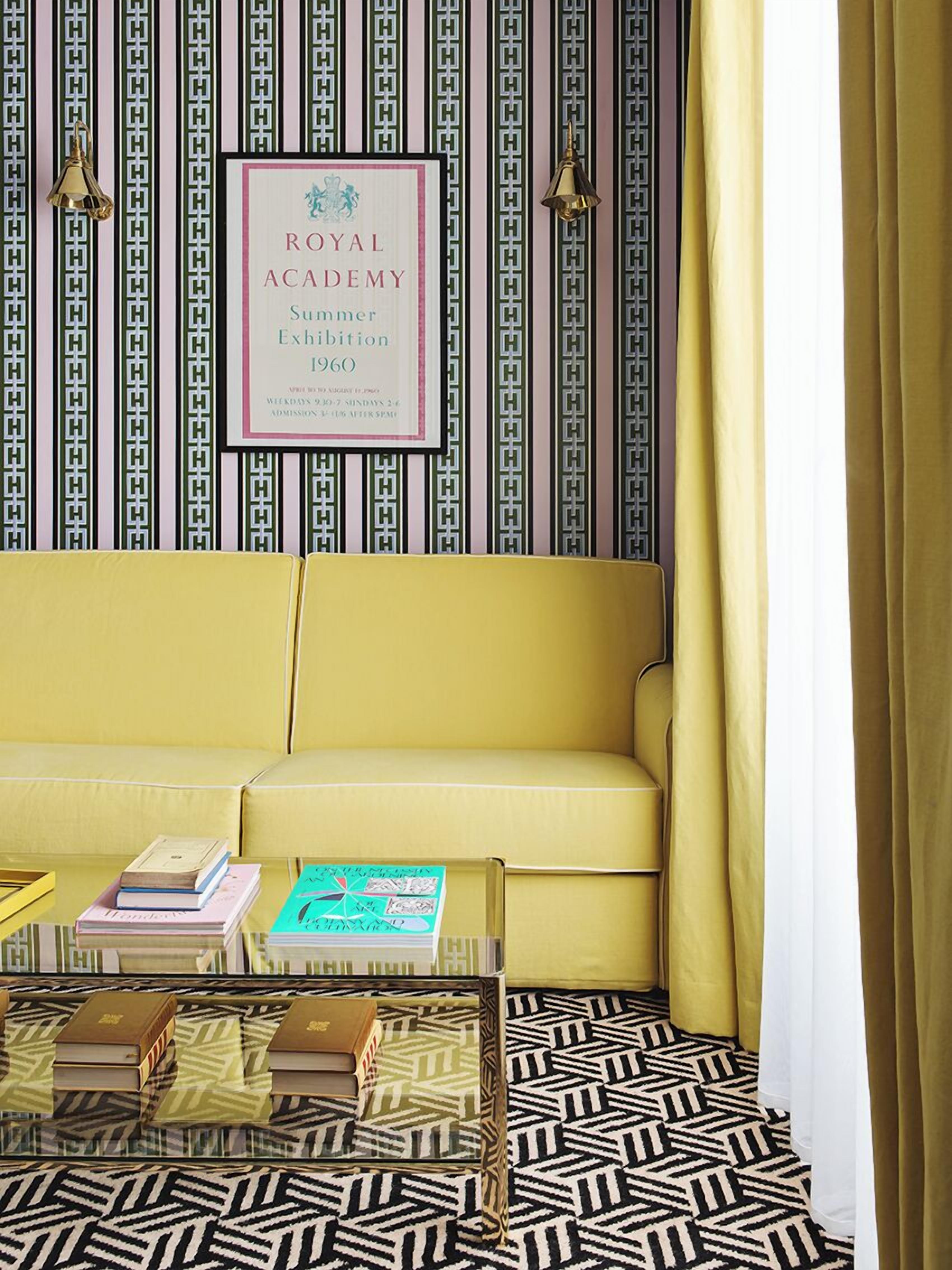 A room with a patterned wallpaper, a framed poster on the wall, a yellow sofa, and a coffee table with books.