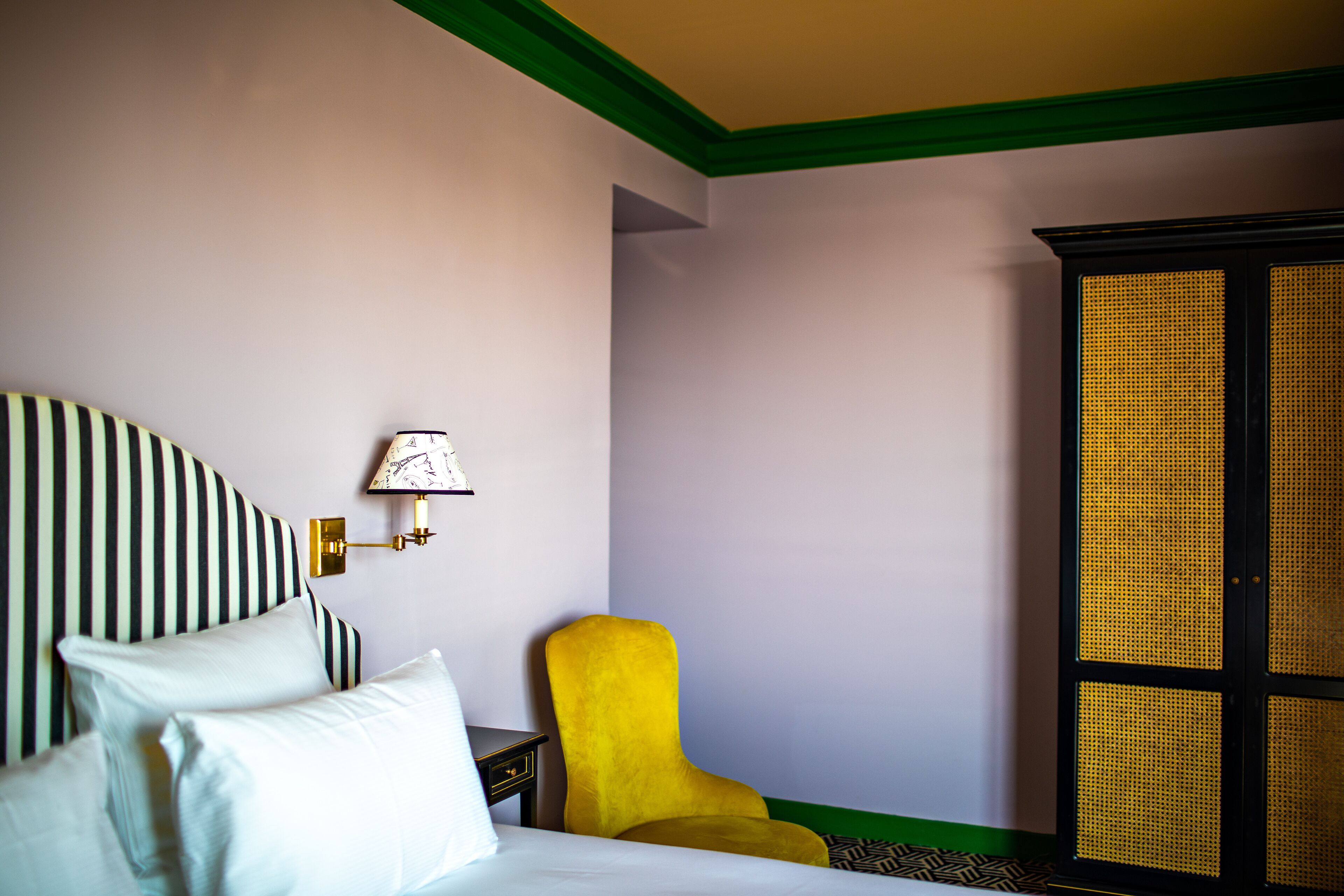 A hotel room with a bed, a yellow armchair, a wall-mounted lamp, and a large window with a woven blind.