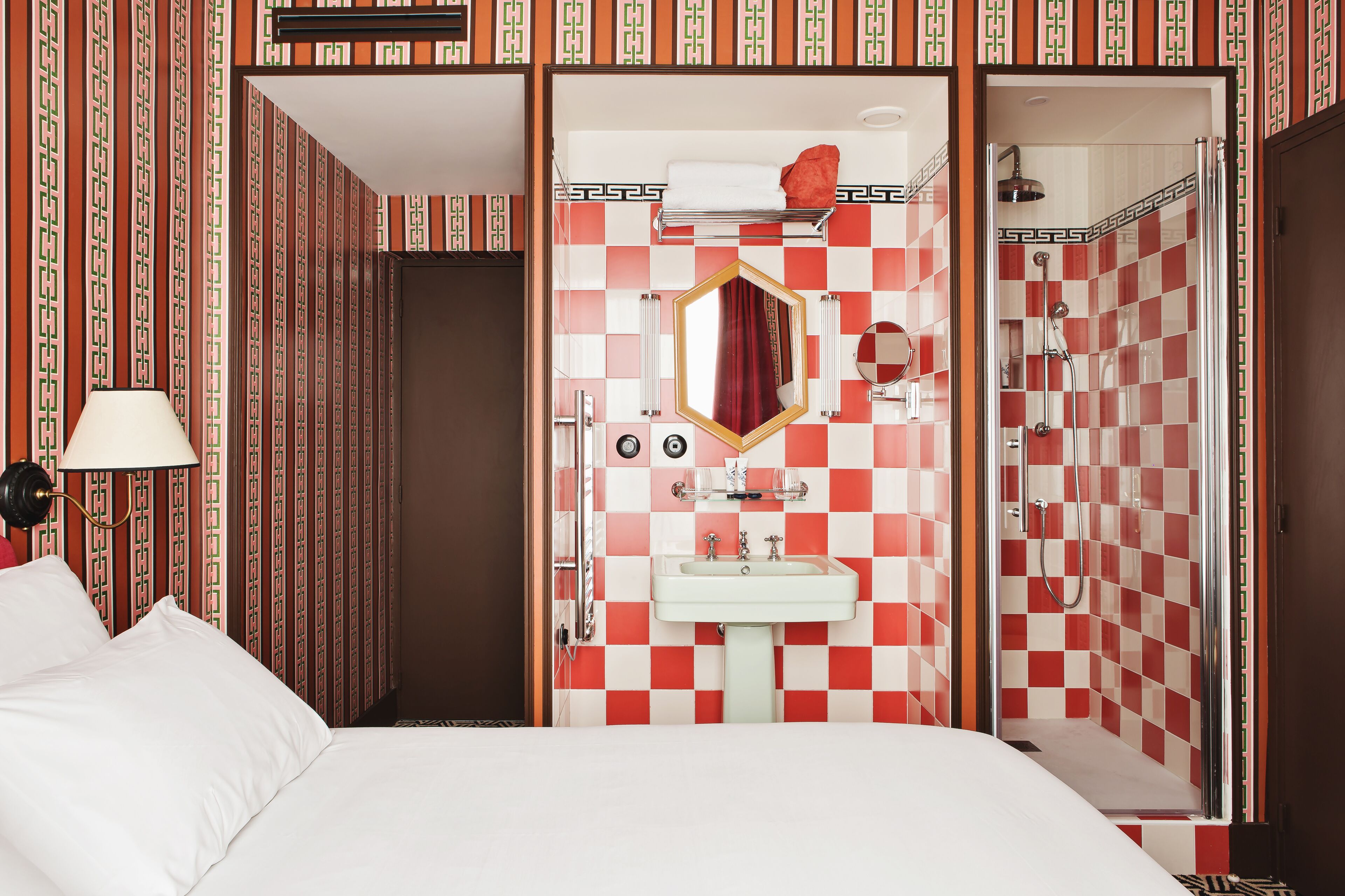 A hotel room with a checkered red and white wall pattern, a white bed, a bedside lamp, a sink, a mirror, and a shower area.