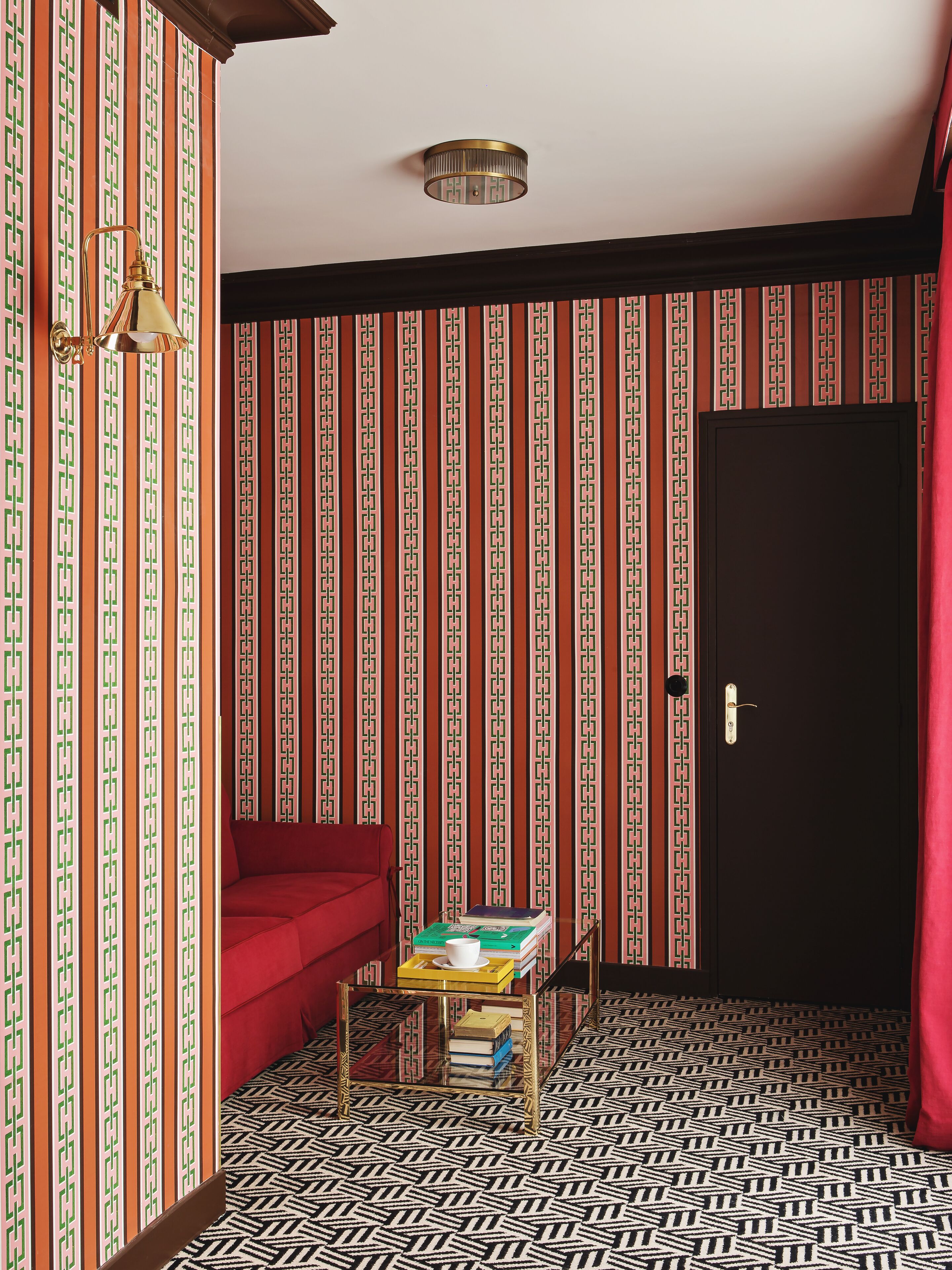 A room with a patterned wallpaper, a red sofa, a small table with books, and a black door.