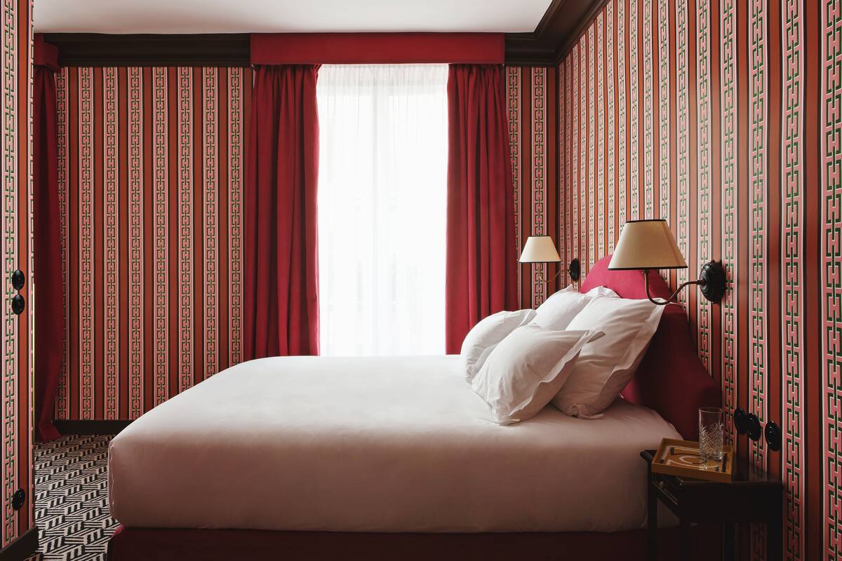 Une chambre d’hôtel avec un grand lit, des rideaux rouges, un papier peint à motifs et une table de chevet avec une lampe et un plateau.