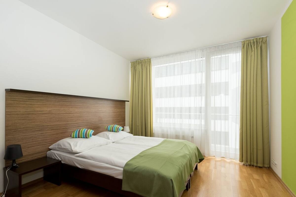 Ein modernes Hotelzimmer mit einem Holzkopfteil, einem Bett mit weißen Leinen und einer grünen Tagesdecke, einem Nachttisch mit einer Lampe sowie durchsichtigen Vorhängen am Fenster.